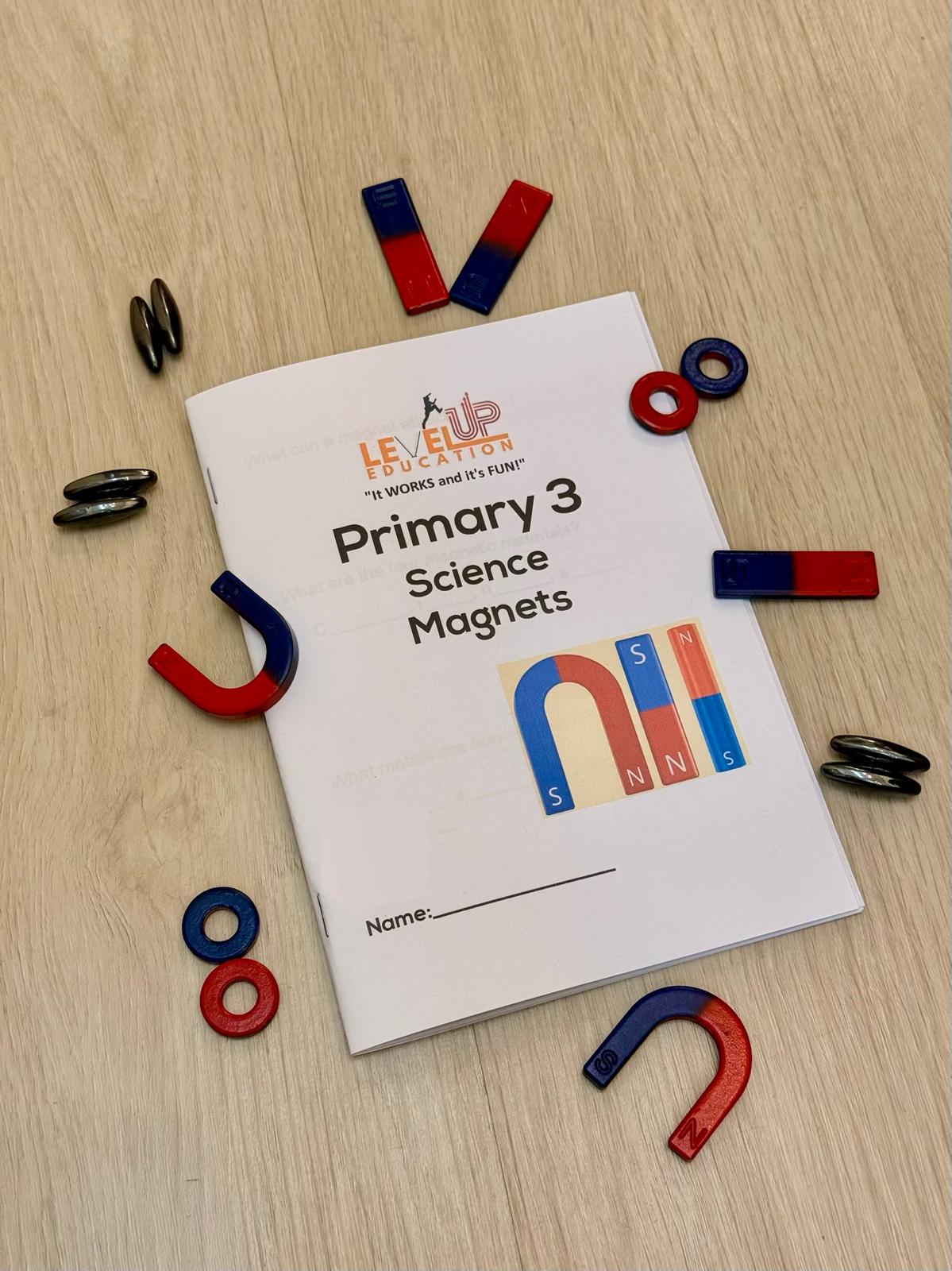 A Primary 3 Science workbook titled "Magnets" by LevelUp Education, surrounded by red and blue horseshoe, bar, and ring magnets on a wooden background